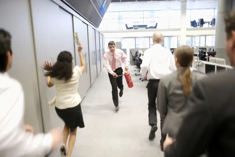 Fotografía de personas corriendo en un piso de empresa mientras un mismo colaborador se acerca al fuego con un extintor para lograr apagar el fuego